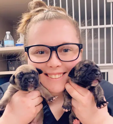Kelsey's staff photo from University West Pet Clinic where she is holding two puppies in front of her face.